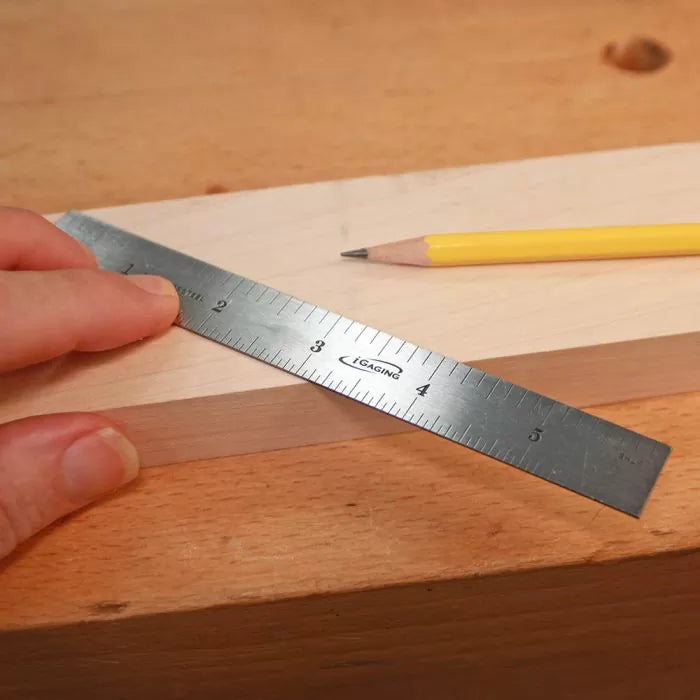 Stainless steel ruler measuring a wooden board with a yellow pencil on a workbench.