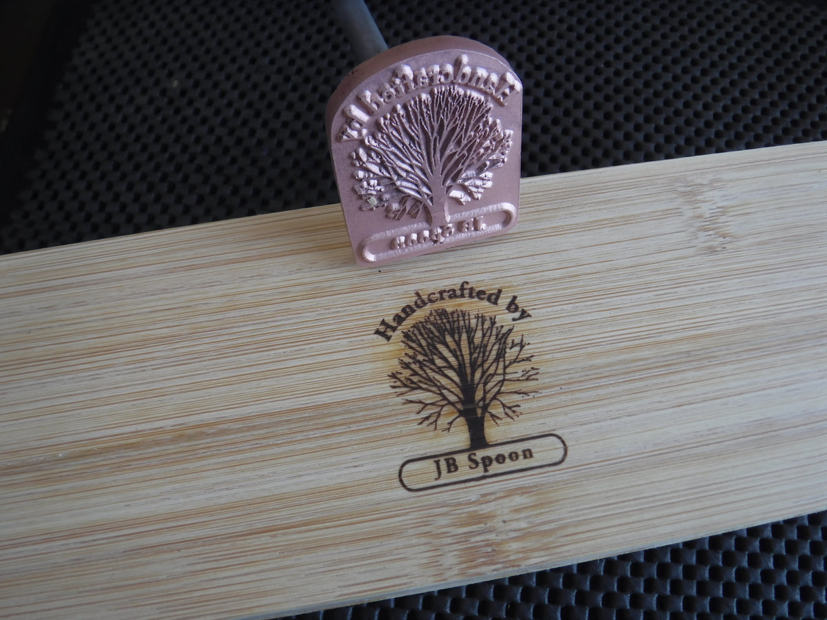 Custom branding iron with tree design marking 'Handcrafted by JB Spoon' on wood.
