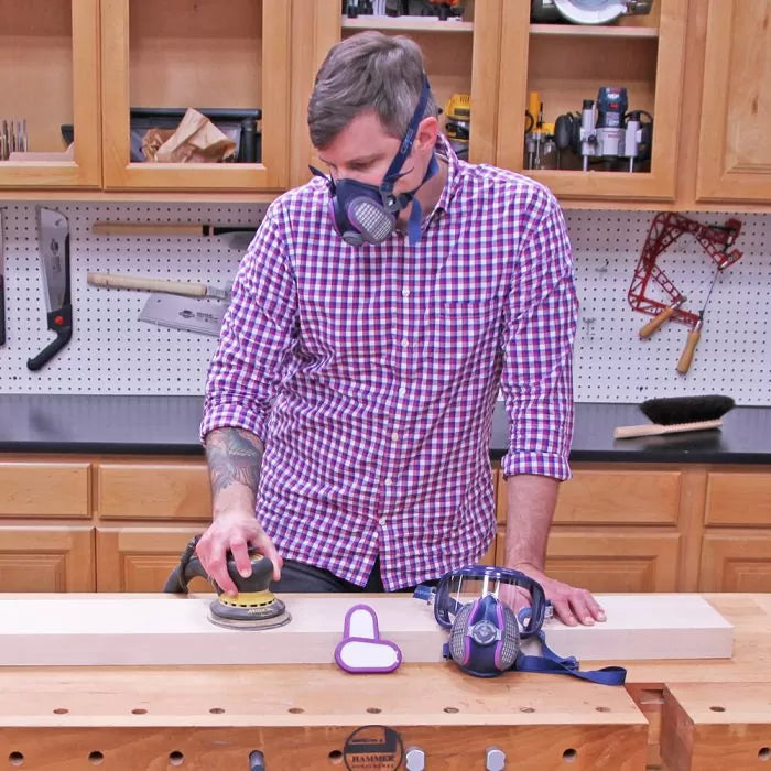 Woodworker sanding board with dust mask in workshop, surrounded by woodworking tools