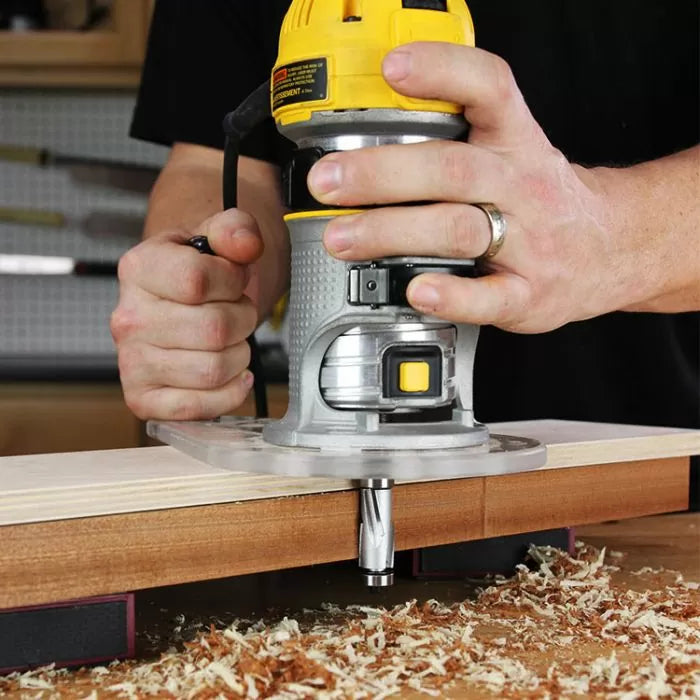 Hands using a router tool to shape wood edge with wood shavings on workbench.