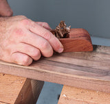 Hand using a wooden hand plane to smooth a hardwood board on a woodworking bench.