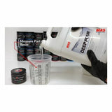 Pouring MAS Deep Pour resin into a measuring cup with containers in the background, woodworking supplies.