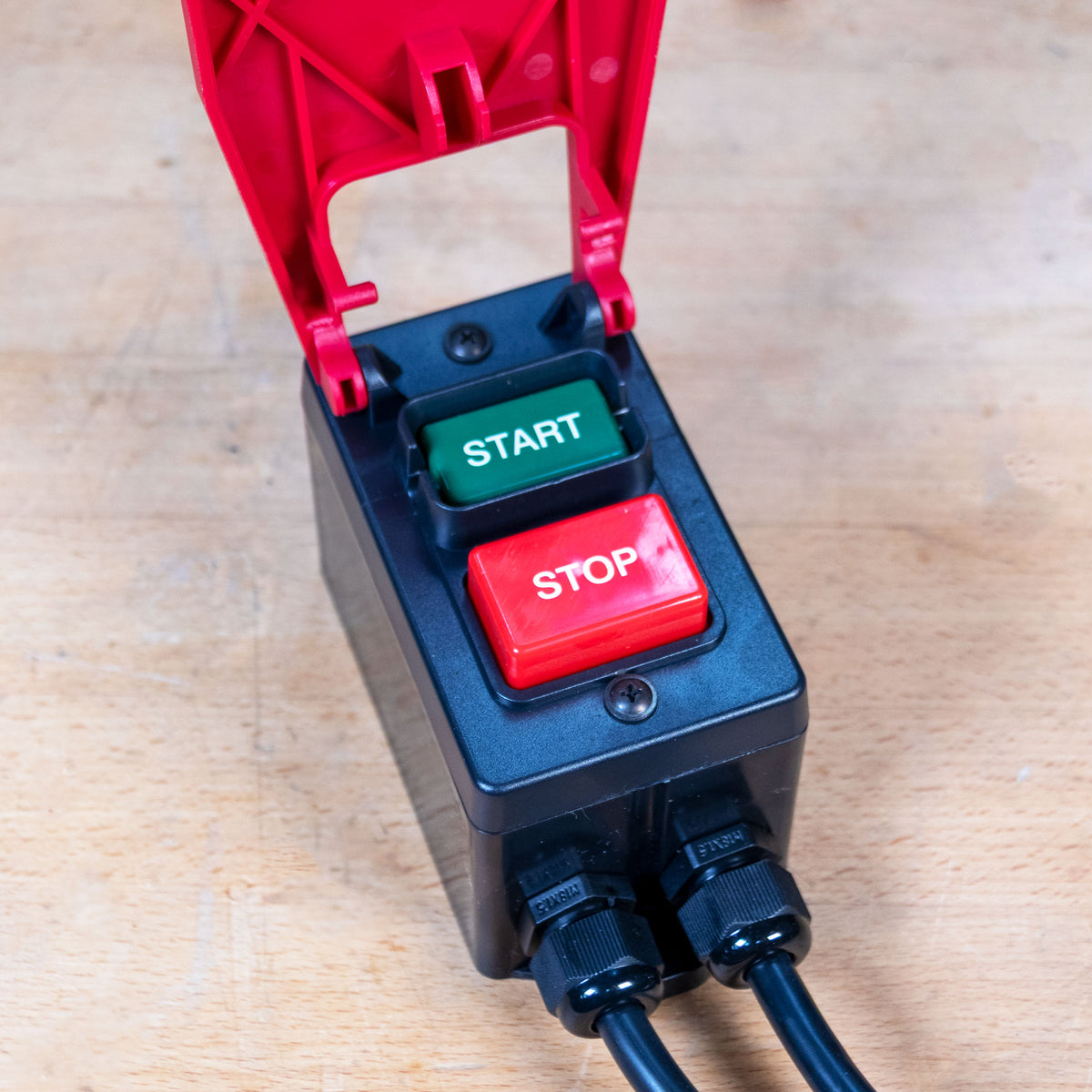 A black electrical control box with a red hinged cover open above it, featuring a green START button and a red STOP button, placed on a light wood surface.