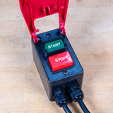 A black electrical control box with a red hinged cover open above it, featuring a green START button and a red STOP button, placed on a light wood surface.
