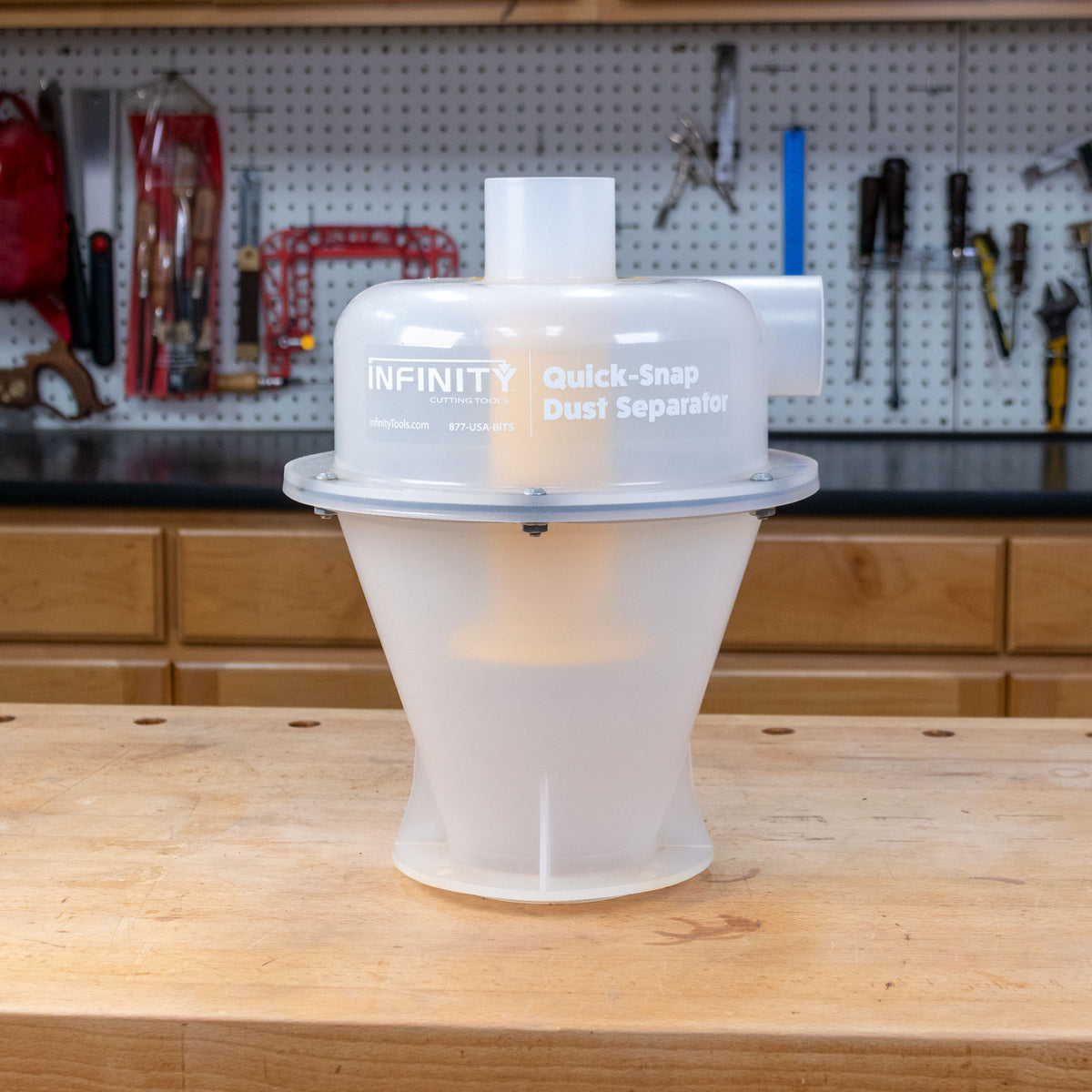 A translucent white plastic dust separator canister with a clear top cap and printed text reading 'INFINITY Quick-Snap Dust Separator,' sitting on a wooden workbench with a workshop pegboard and tools in the background.