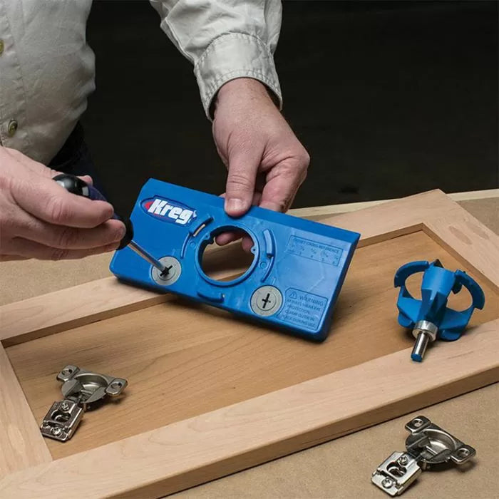 Person using a blue Kreg cabinet hardware jig on a wooden cabinet door with hinges and a router bit nearby.