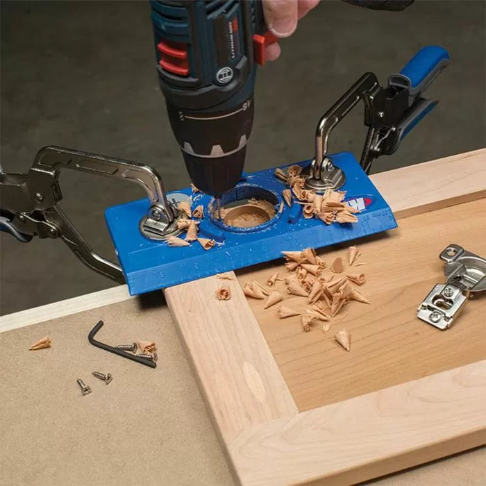 Drill guide jig clamped on cabinet door for hinge installation with wood shavings