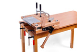 A wooden tabletop clamp fixture with metal C-clamps and a wooden miter guide, shown mounted on a wooden workbench against a white background.