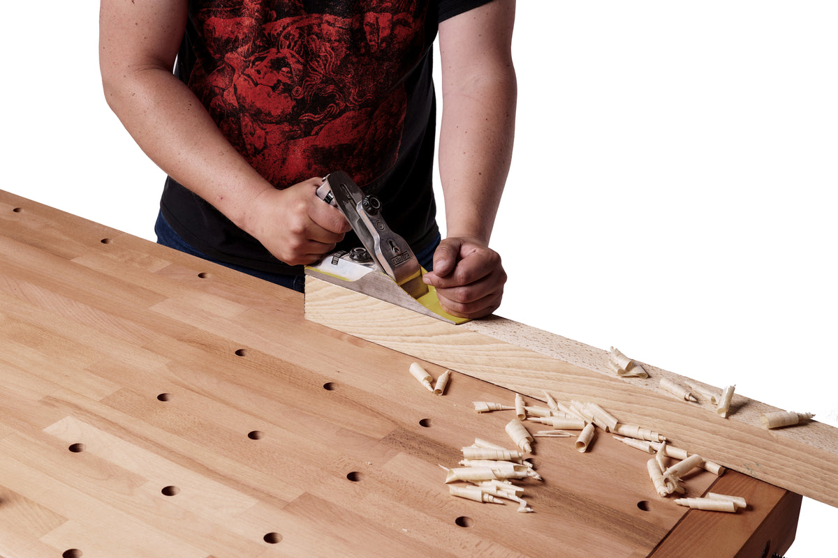 A person using a hand plane to shave wood shavings from a light-colored wooden board on a workbench, with wood shavings scattered around on a white background.