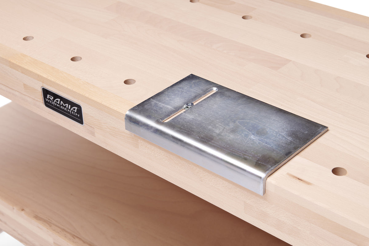 A close-up of a silver metal workbench vise plate mounted on a light-colored wood workbench. A black RAMA brand label is visible on the left side of the bench.