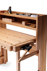 A light natural wood workbench with a peg-hole slatted desktop, open shelving, and a black-and-tan manual clamp mechanism on the right side, shown on a white background.