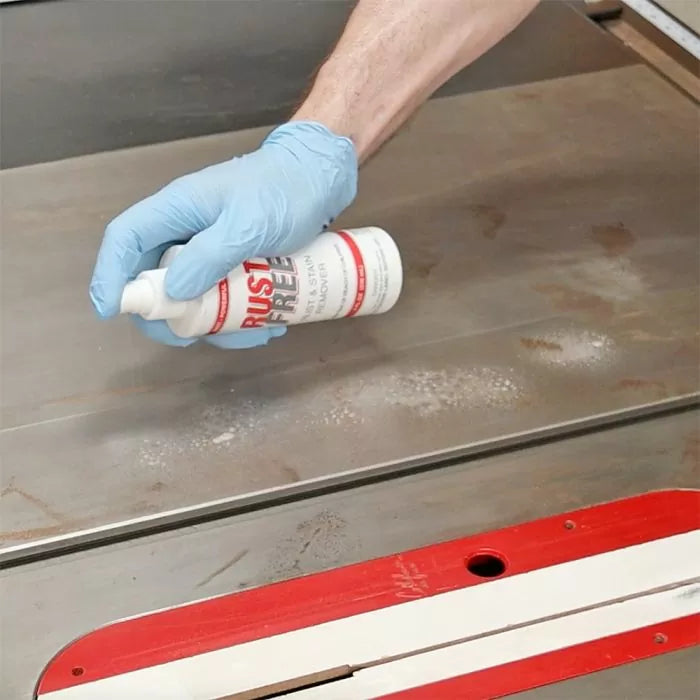 Gloved hand applying rust remover spray to a table saw top in a woodworking shop.